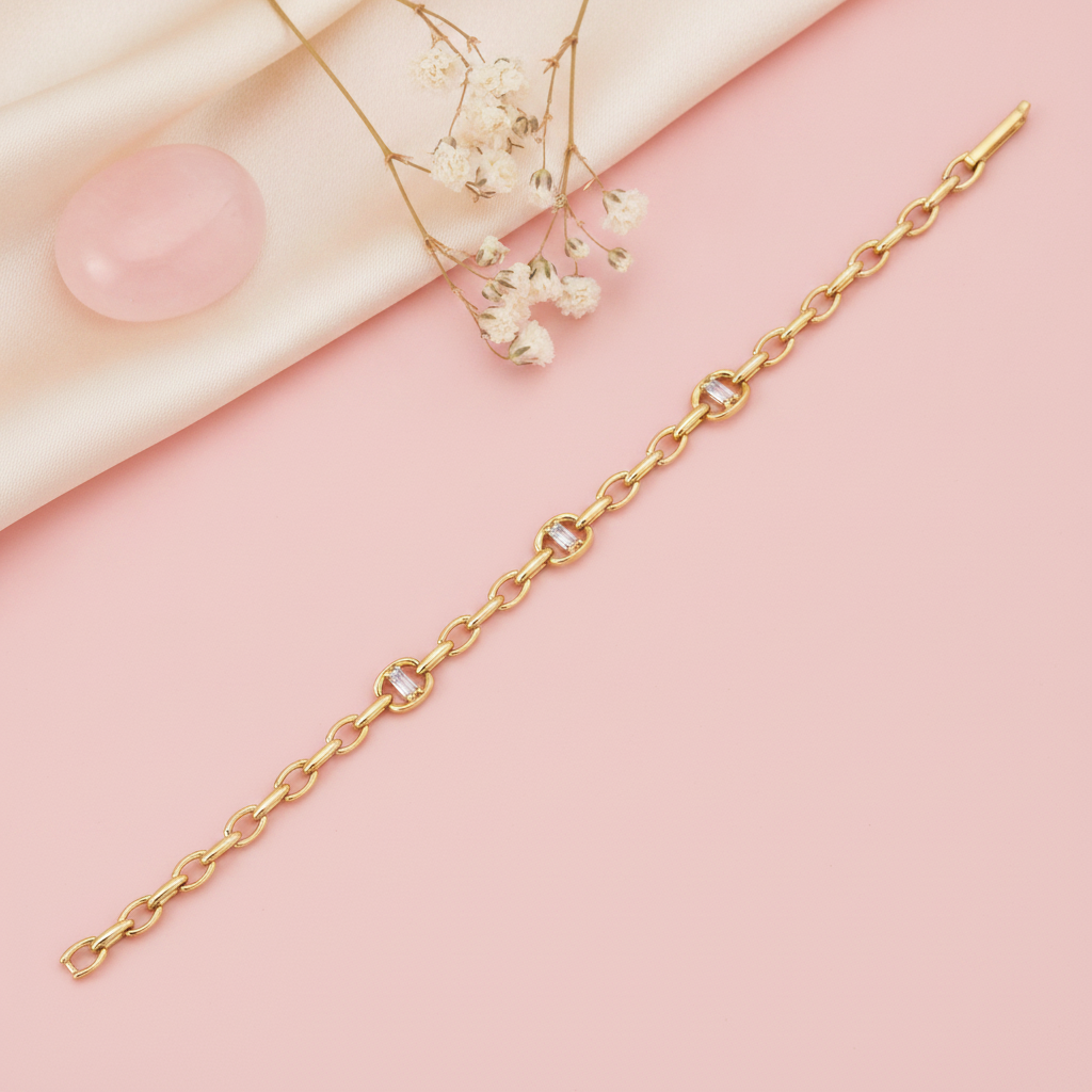gold open-link chain bracelet with framed baguette stones laid diagonally on pink background with dried flowers