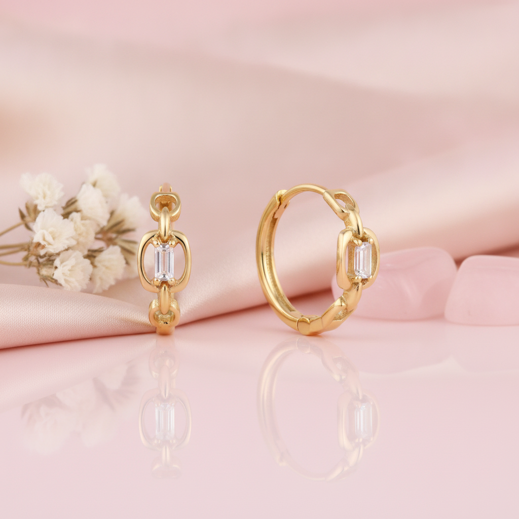 gold chain-link huggie hoop earrings with a baguette stone, one shown in profile and one facing forward on pink satin