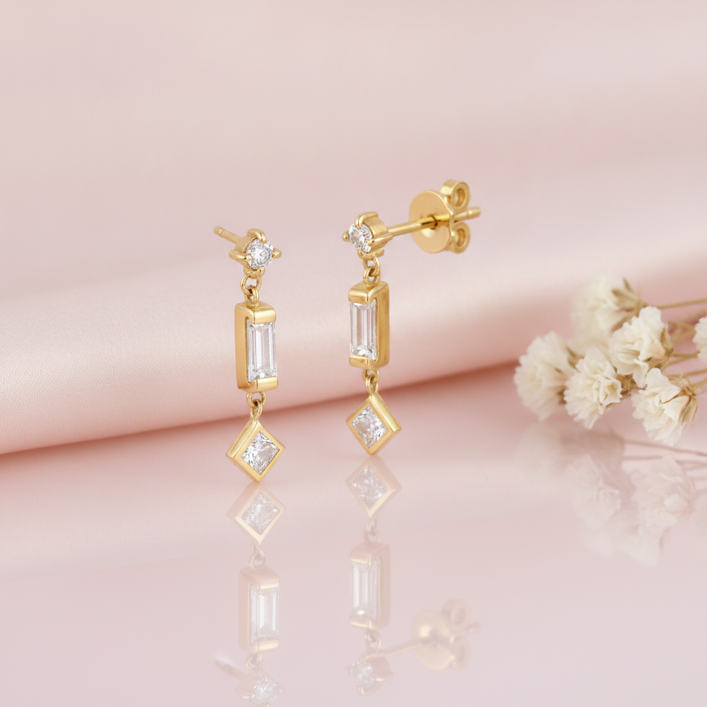 gold articulated drop earrings with a round stud, baguette stone, and square stone drop on pink satin with dried flowers