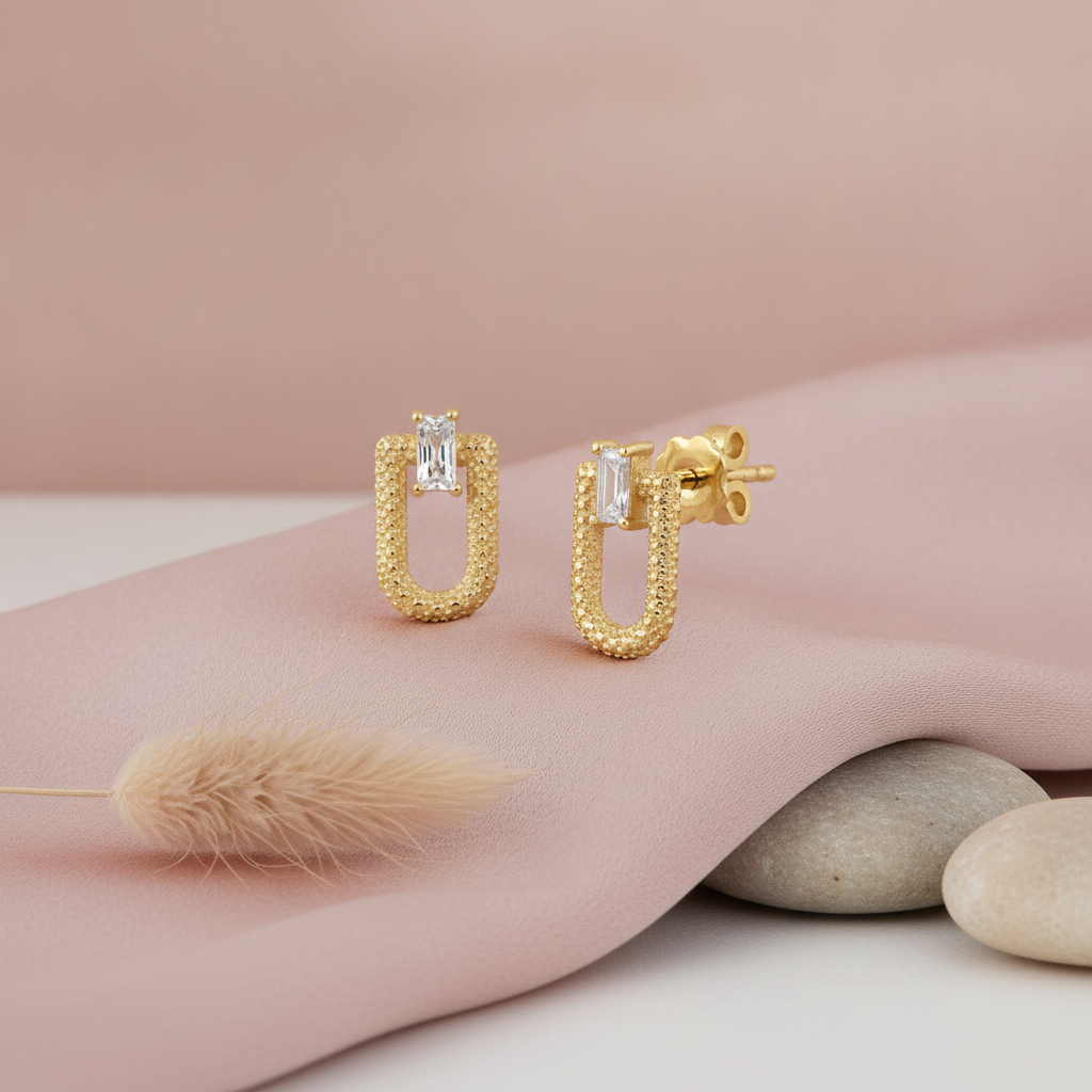 gold textured open-link stud earrings with a baguette stone, on pink satin with dried bunny tail grass and pebbles