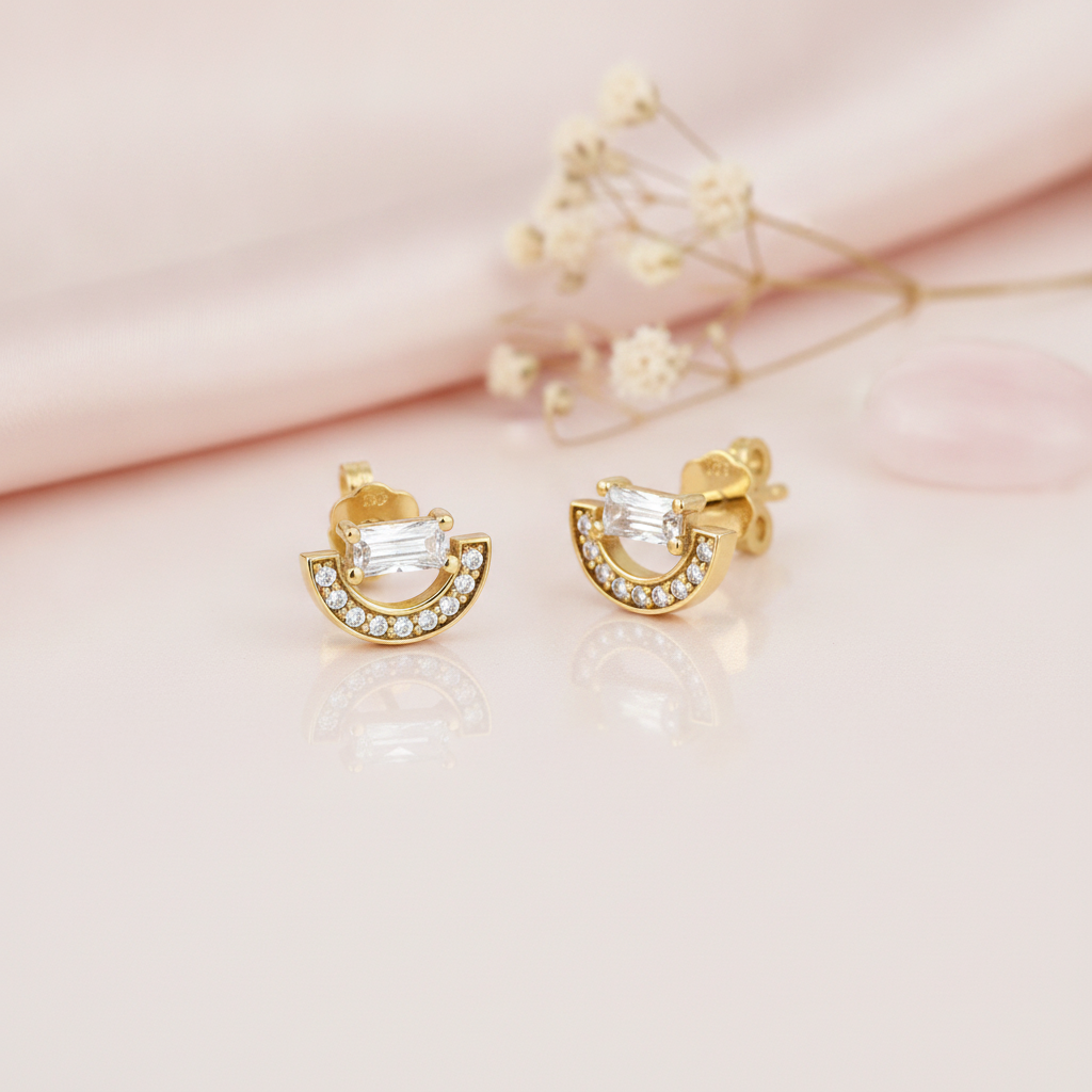 gold stud earrings with a pave crescent arc topped by a horizontal baguette stone, on pink satin with dried flowers and a rose quartz stone