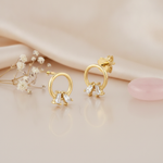 gold open circle stud earrings with two angled baguette stones at the base, on pink satin with dried flowers and a pink stone