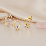 gold open circle stud earrings with two angled baguette stones at the base, on pink satin with dried flowers and a pink stone