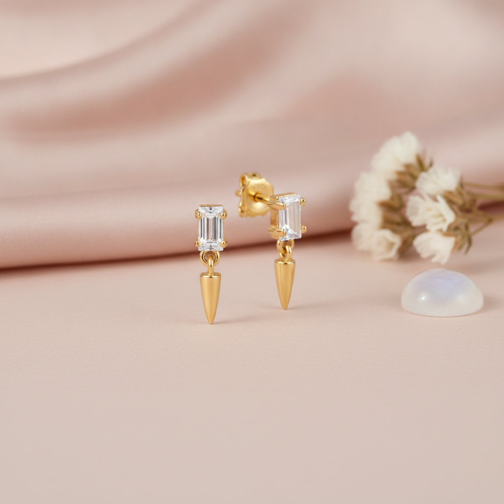 gold stud earrings with a baguette stone and a dangling gold spike drop, on pink satin with dried flowers and a moonstone cabochon