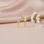 gold stud earrings with a baguette stone and a dangling gold spike drop, on pink satin with dried flowers and a moonstone cabochon