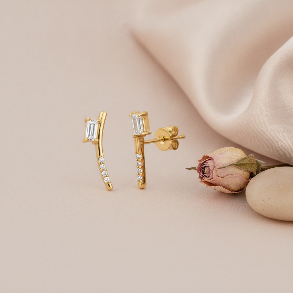 gold curved stud earrings with a baguette stone and round accent stones, on blush background with a dried rose and pebbles