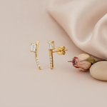 gold curved stud earrings with a baguette stone and round accent stones, on blush background with a dried rose and pebbles