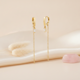 gold huggie hoop earrings with a baguette stone and long beaded chain drop, on cream background with dried bunny tail grass and a rose quartz stone