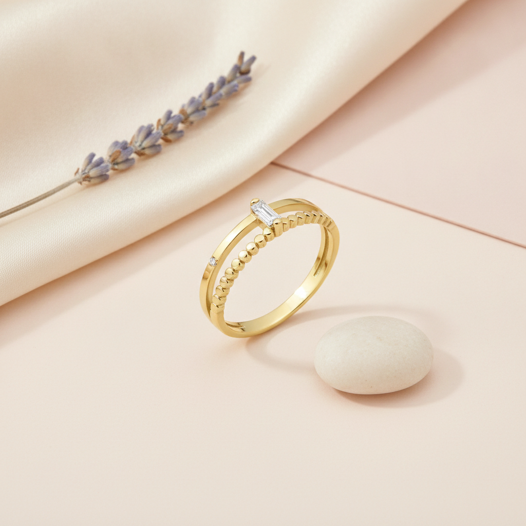 gold dual-band ring, one polished and one beaded, with a baguette stone and a round accent stone, on cream satin with a lavender sprig and pebble