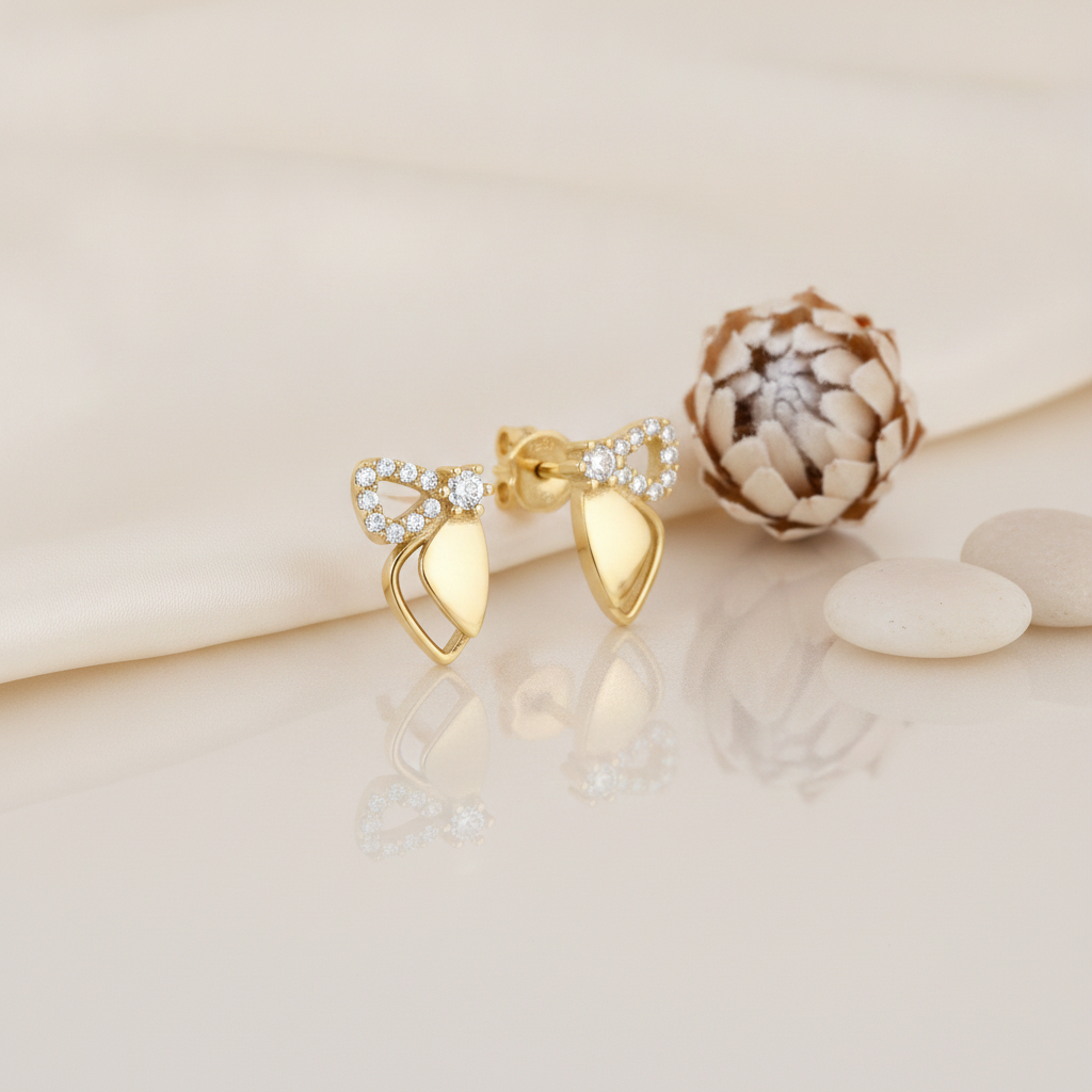 gold bow stud earrings with pave stones and a polished gold petal drop, on cream satin with a pinecone and pebbles