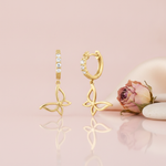 gold hoop earrings set with round stones and dangling openwork butterfly charms, on pink background with a dried rose and pebbles