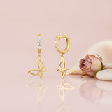 gold hoop earrings set with round stones and dangling openwork butterfly charms, on pink background with a dried rose and pebbles