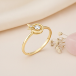 gold ring with a round clear stone halo and butterfly accent displayed on cream fabric with dried flowers and a smooth stone