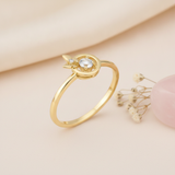 gold ring with a round clear stone halo and butterfly accent displayed on cream fabric with dried flowers and a smooth stone
