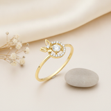 gold ring with a pave stone halo and side butterfly motif displayed on cream fabric with dried flowers and a smooth gray stone