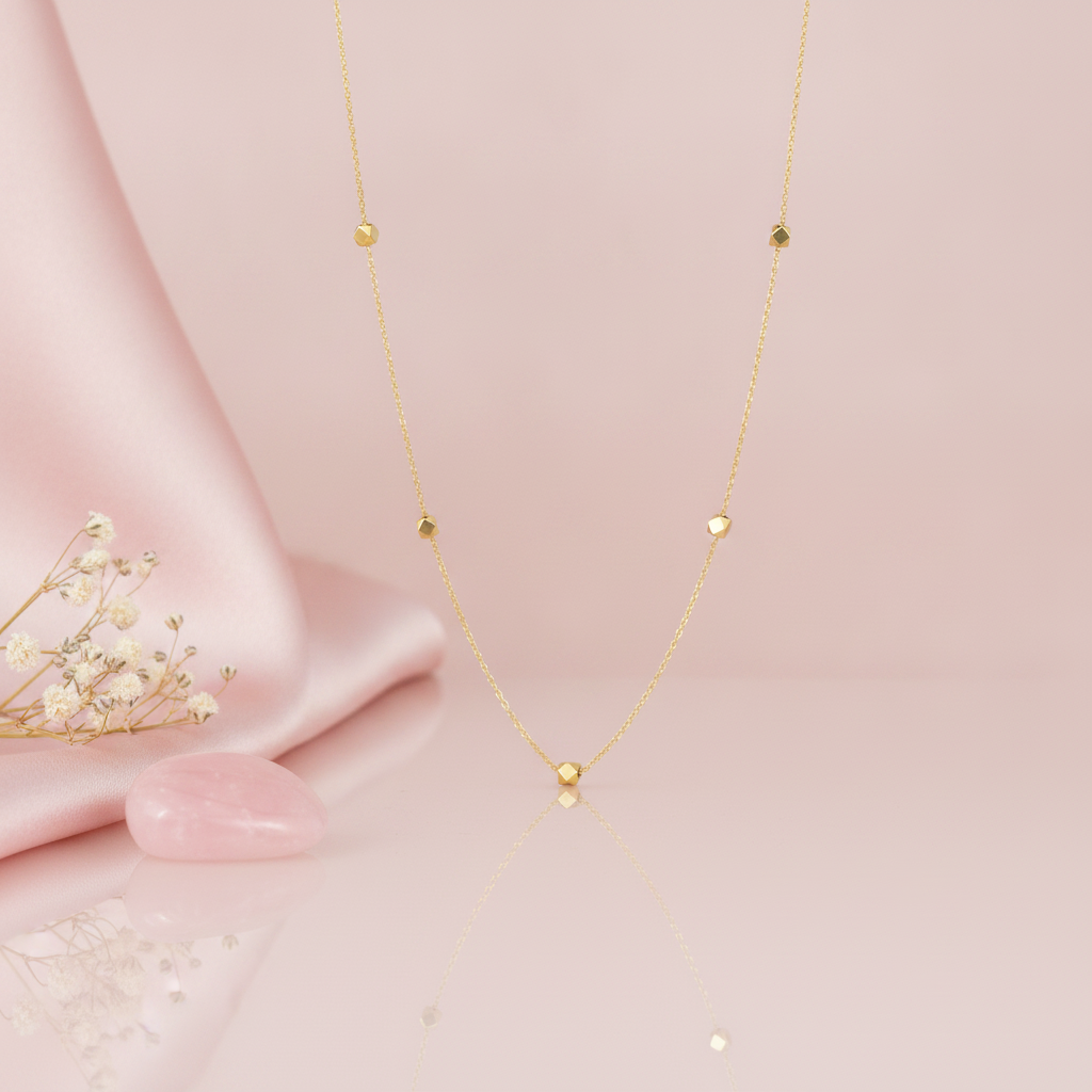 gold station necklace with faceted nuggets hanging against a blush pink background with dried flowers and a pink stone