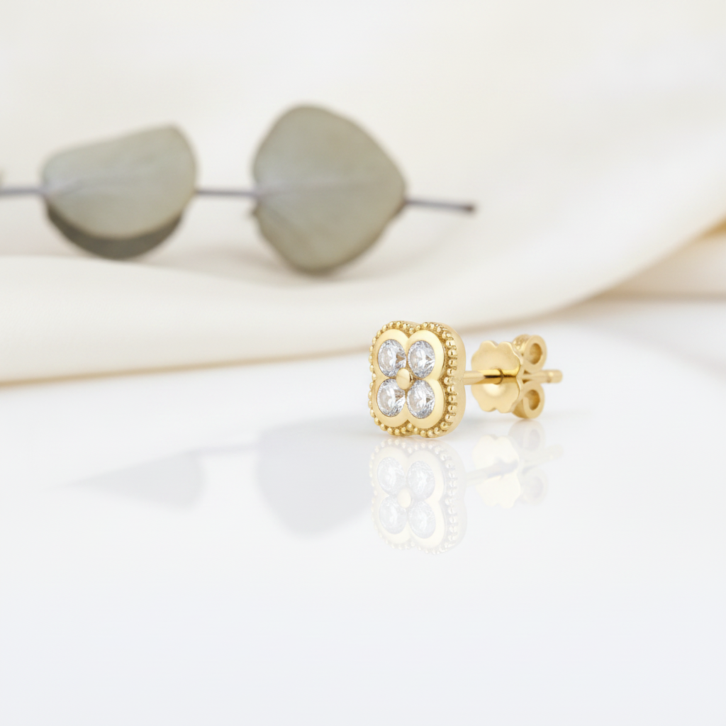 gold clover stud earring with White CZ stones on a reflective white surface with a blurred eucalyptus leaf in the background