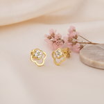 pair of gold open clover stud earrings with White CZ stones, styled with pink flowers on a cream background