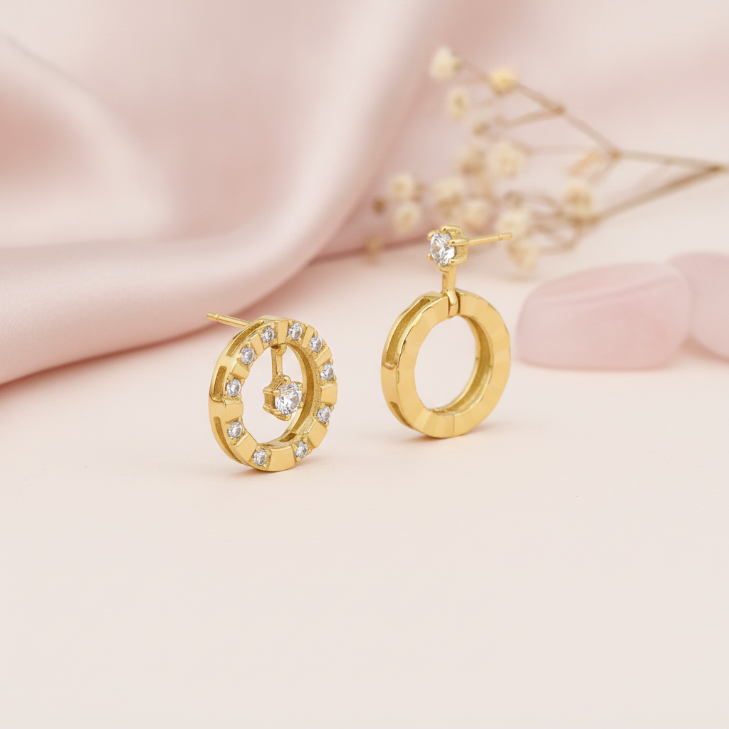 Asymmetric gold earrings with a pave circle and suspended stone beside a faceted hoop, styled on pink satin with dried flowers