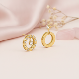 Asymmetric gold earrings with a pave circle and suspended stone beside a faceted hoop, styled on pink satin with dried flowers