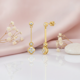 Gold drop earrings with a textured bar and quatrefoil stone cluster, shown front and back on pink satin with dried flowers