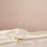 Gold station necklace with five clover motifs and an open outline drop pendant on a pink background with dried flowers