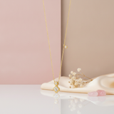 Gold necklace with a four-petal clover pendant, a bezel-set accent stone, and an off-center charm on a pink background with a dried rose