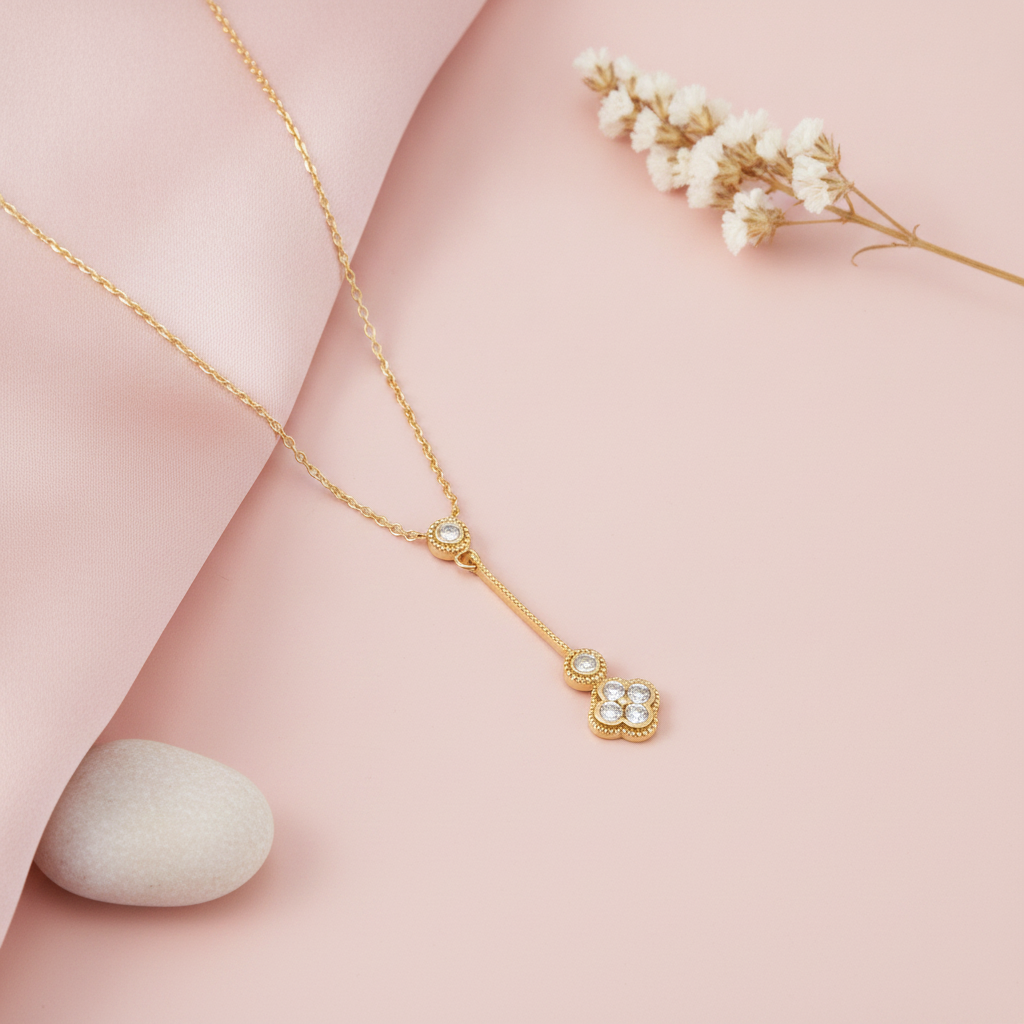 Gold Y-necklace with a textured vertical bar and a clover-shaped stone drop on a pink background with dried flowers