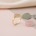 yellow gold ring with a circular cluster of round clear stones, styled on a pink and taupe background with a eucalyptus leaf sprig and a rose quartz stone