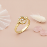 yellow gold ring with an open circular halo of pave stones and a center stone, styled on a blush pink background with tulle, a dried pampas plume, and rose quartz stones