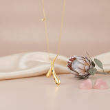 yellow gold crossed teardrop pendant necklace with blue and clear accents, styled on cream silk with a dried protea flower and rose quartz stones