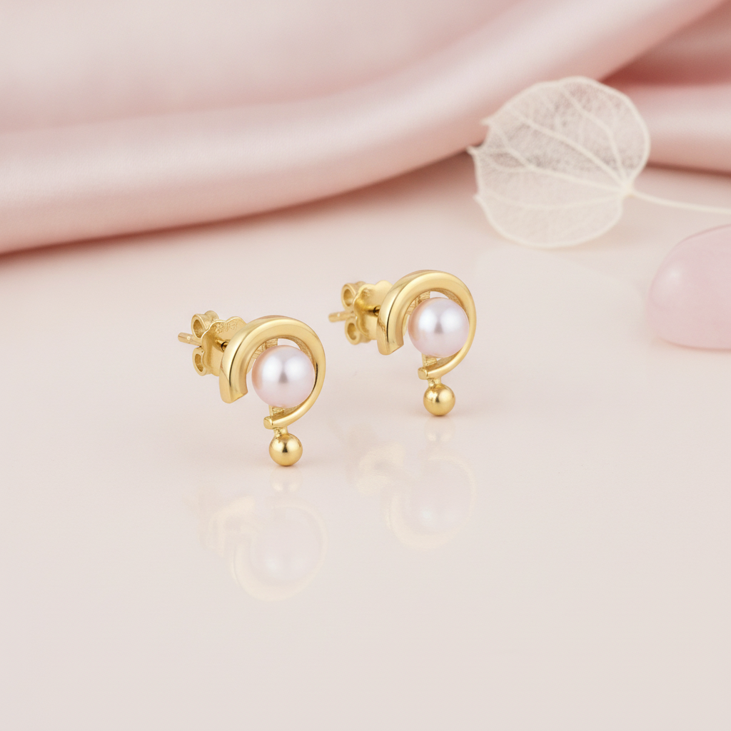 yellow gold arc stud earrings with a pearl, styled on a pink background with a dried leaf and a rose quartz stone