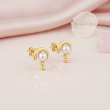 yellow gold arc stud earrings with a pearl, styled on a pink background with a dried leaf and a rose quartz stone