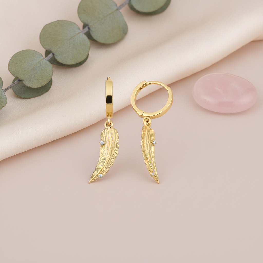 yellow gold huggie hoop earrings with textured feather drops, styled on cream fabric with eucalyptus leaves and a rose quartz stone