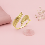 yellow gold open wrap ring with two textured feathers and accent stones, styled on a pink background with dried baby's breath and a rose quartz stone