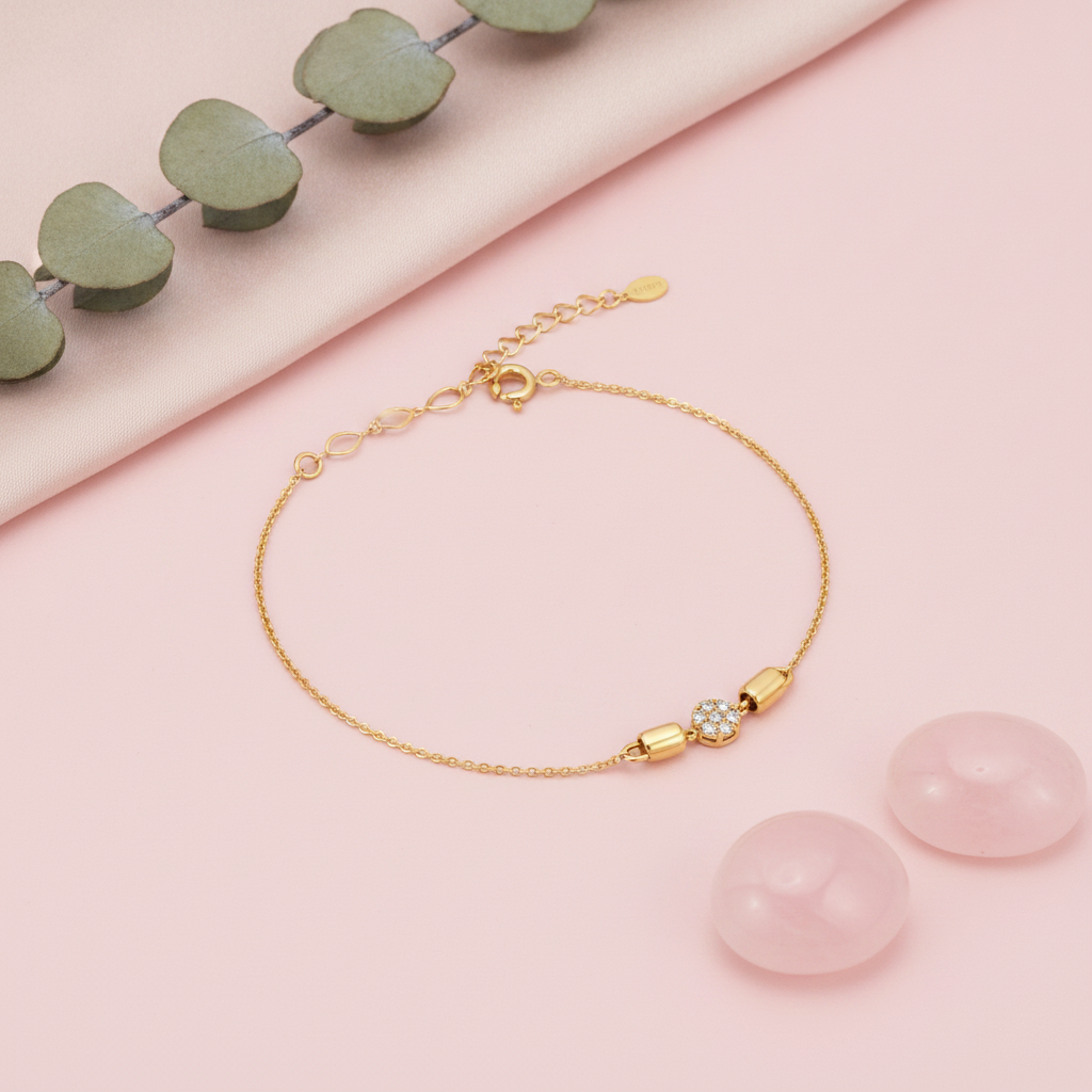 gold bracelet with a pave disc charm flanked by polished capsule beads, shown on pink fabric with eucalyptus leaves and rose quartz stones