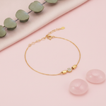 gold bracelet with a pave disc charm flanked by polished capsule beads, shown on pink fabric with eucalyptus leaves and rose quartz stones