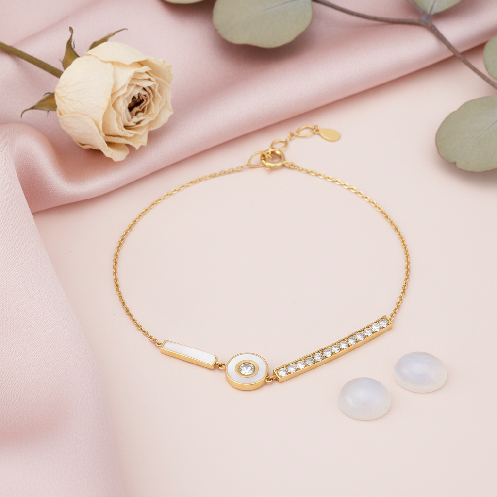 gold bracelet with a bar of clear stones and a white-inlay disc charm, shown on pink fabric with a dried rose and eucalyptus leaves