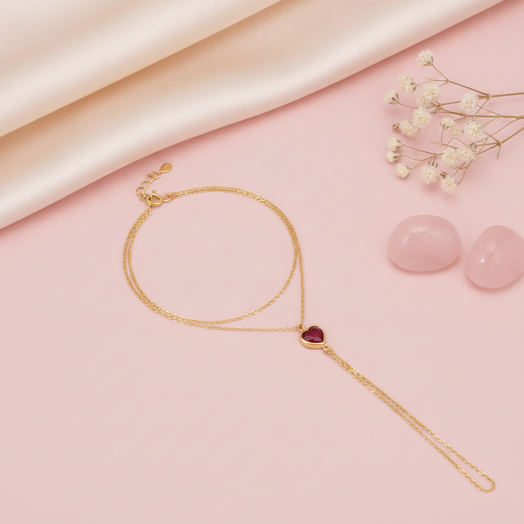 gold hand chain bracelet with a heart-shaped red stone charm, shown on pink fabric with baby's breath and rose quartz stones