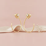 pair of gold hoop earrings topped with a heart motif and a small stone, shown on pink fabric with dried baby's breath