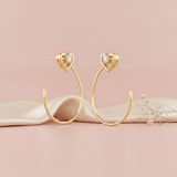 pair of gold hoop earrings topped with a heart motif and a small stone, shown on pink fabric with dried baby's breath