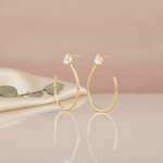 pair of open hoop earrings topped with a pear-cut stone, shown on a reflective surface with eucalyptus and cream fabric