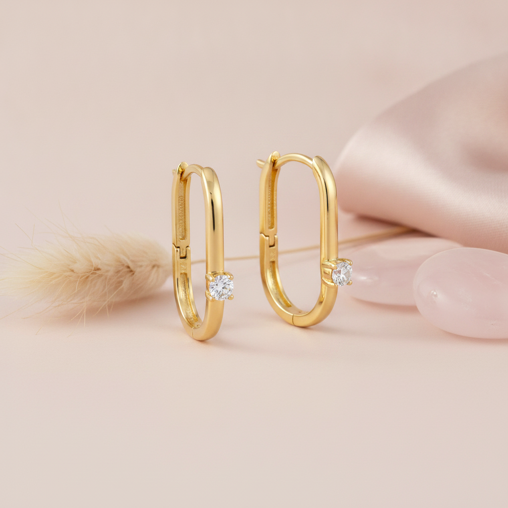 gold elongated oval hoop earrings with a single round White CZ stone on a blush background with a dried grass sprig