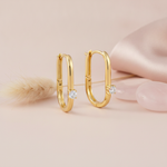 gold elongated oval hoop earrings with a single round White CZ stone on a blush background with a dried grass sprig