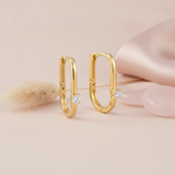 gold elongated oval hoop earrings with a single round White CZ stone on a blush background with a dried grass sprig