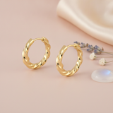 polished gold twisted huggie hoop earrings on a blush background with lavender sprigs and a translucent stone