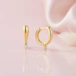 gold tapered drop earring and huggie hoop with a single round White CZ stone on a pink satin background with dried grass