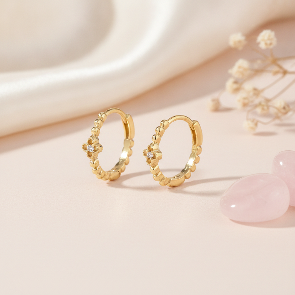 gold beaded huggie hoop earrings with a quatrefoil motif and a White CZ stone on a blush background with rose quartz pebbles