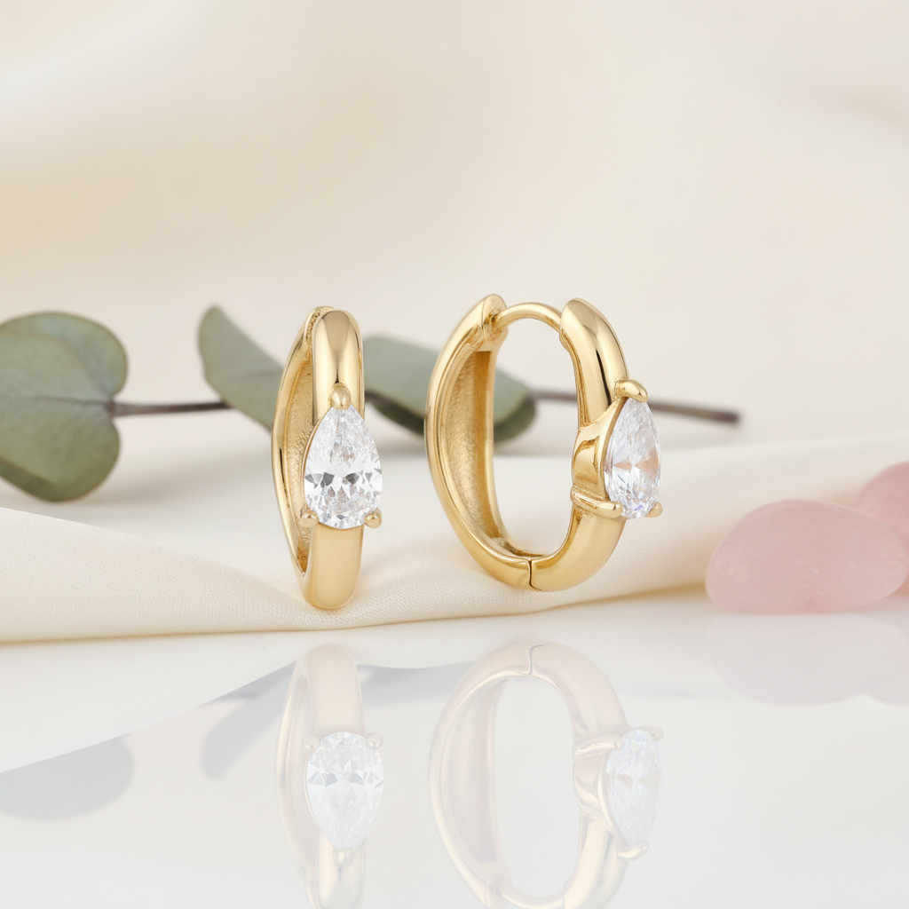 solid gold huggie hoop earrings with a single pear-cut stone on a cream background with eucalyptus leaves and rose quartz pebbles