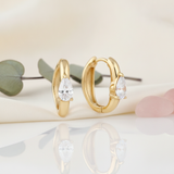 solid gold huggie hoop earrings with a single pear-cut stone on a cream background with eucalyptus leaves and rose quartz pebbles